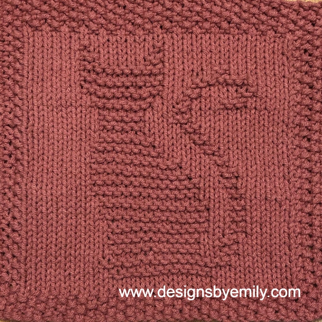 Cat Knit Dishcloth: A handmade knit dishcloth square in dark pink yarn featuring a cat in the center sitting tall with its tail curling up and to the right. The design is created in garter stitch using knit and purl stitches on a stockinette background.